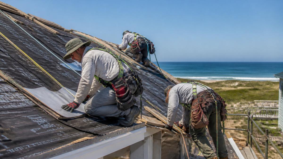 A roofing crew in safety harnesses installing underlayment and flashing on a coastal home roof, with the ocean visible in the background under a clear sky